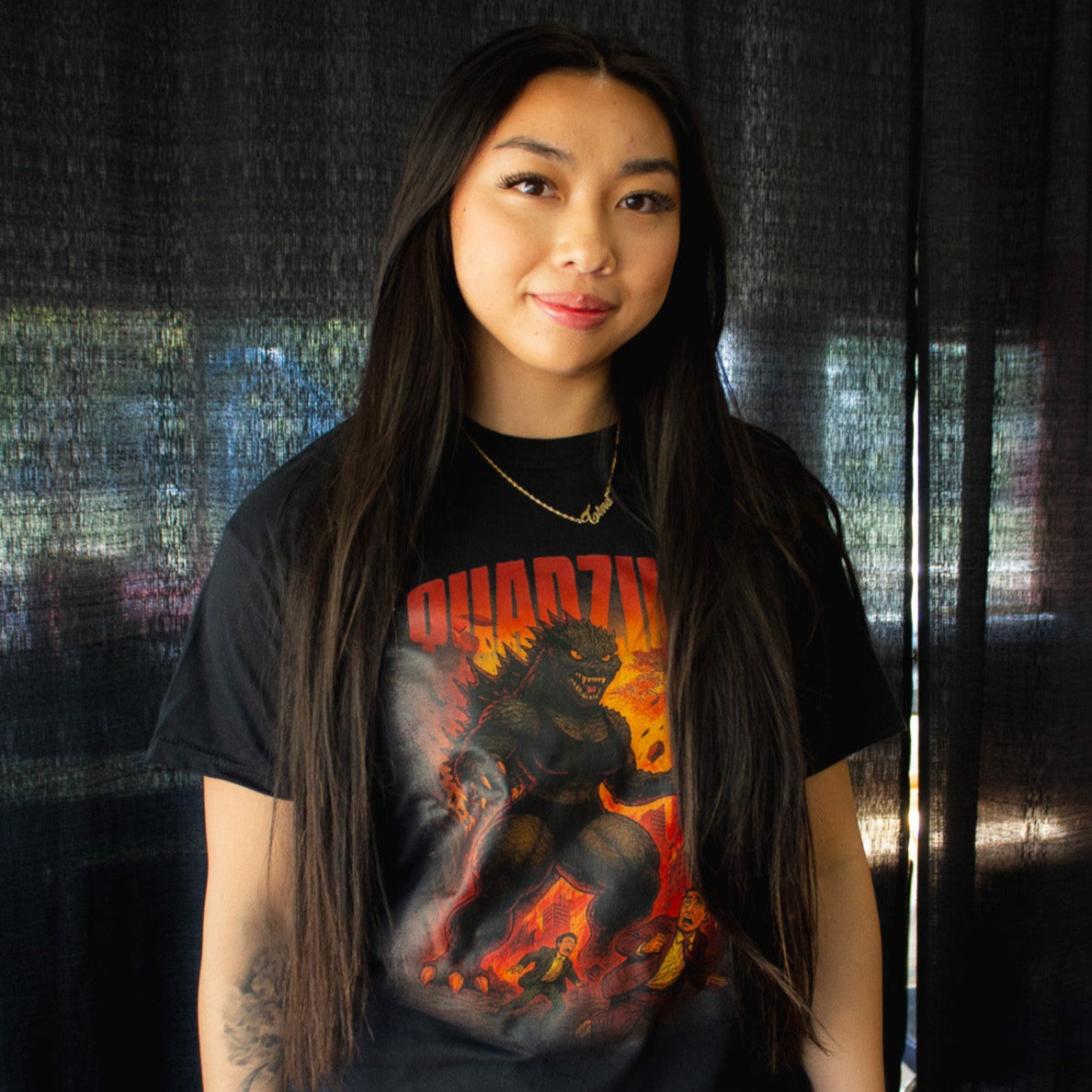 Person wearing a black t-shirt with a graphic design in front of a dark curtain.