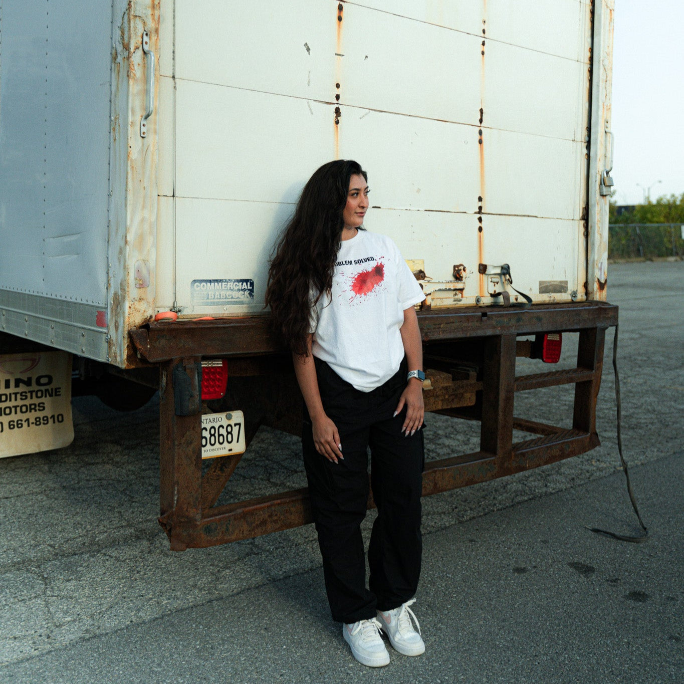 Person leaning against a large white truck with visible rust, standing on a concrete surface. dexter inspired shirt