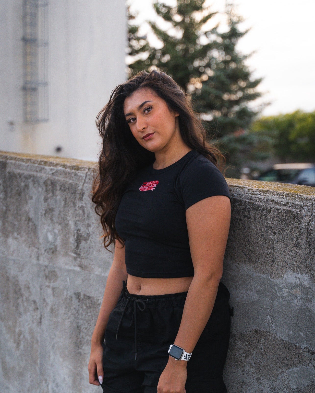 Woman wearing a black crop top and pants leaning against a concrete wall with trees and buildings in the background.
