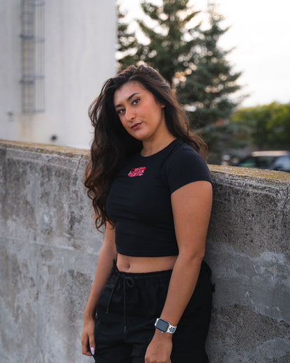 Woman wearing a black crop top and pants leaning against a concrete wall with trees and buildings in the background.