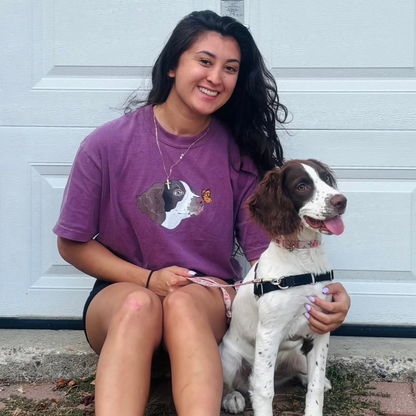 A woman sitting outdoors wearing a purple t-shirt with a custom pet portrait of a dog on it, alongside the dog that is sitting on her lap.